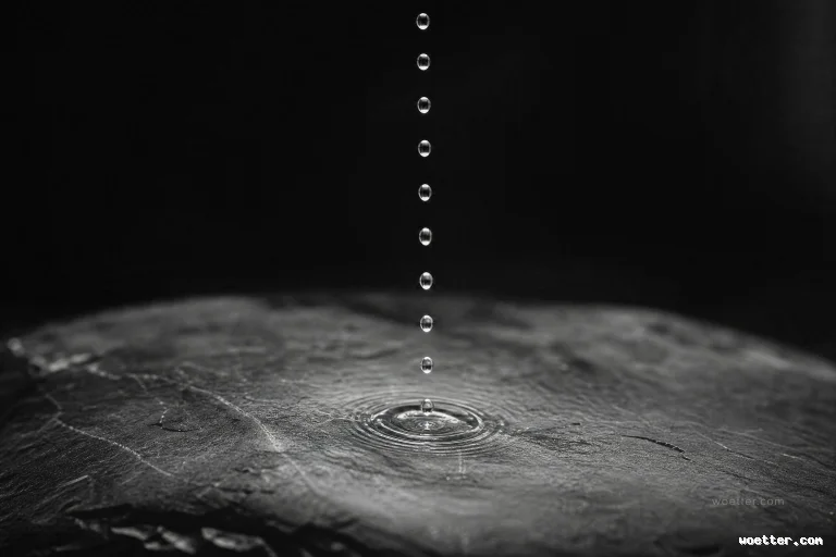 High-contrast black and white photograph showing a perfectly aligned vertical line of water droplets falling toward a dark, textured stone surface. The bottom droplet has just impacted the stone, creating small, concentric circular ripples in a shallow layer of water. The background is completely pitch black, isolating the illuminated droplets and the raw details of the rock in a minimalist atmosphere.
