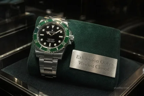 Um relógio de luxo Rolex Submariner de aço prateado, com mostrador preto e aro verde, exibido sobre uma almofada de veludo verde-escuro dentro de uma vitrine. Ao lado direito do relógio, uma pequena placa de metal prateado exibe a inscrição gravada "Exhibition Only - Waitlist Closed". A iluminação focada destaca o relógio e a placa, enquanto o fundo escuro e reflexivo cria uma atmosfera de alta joalheria.