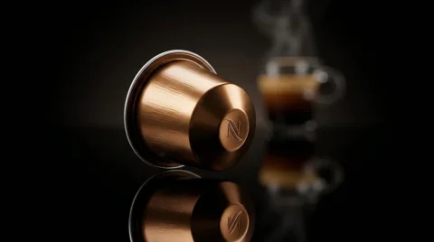 A copper-colored metallic coffee capsule lying on its side on a highly reflective black surface, displaying a sharp reflection. The capsule features an elegant brushed texture and an embossed "N" on its base. In the softly blurred background, a clear glass cup holds steaming espresso, completing a dark, sophisticated setting with dramatic lighting.